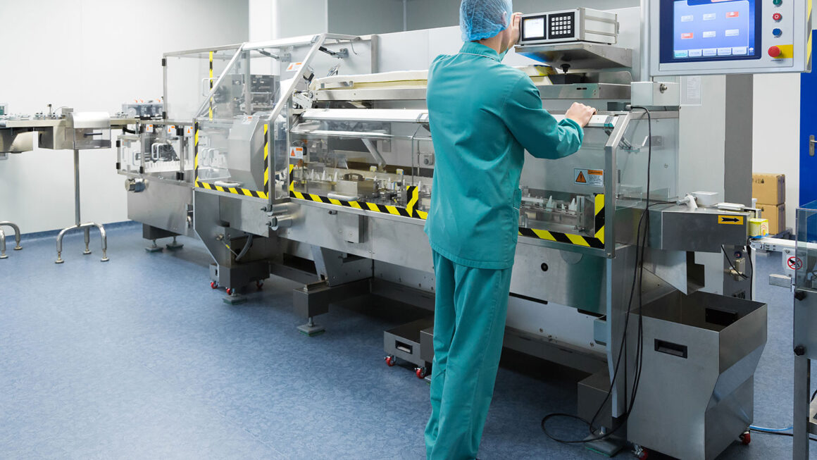 İlaç fabrikasında hijyenik kıyafetli operatör, üretim bandında makineyi kontrol ediyor.